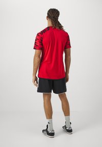 Maglietta atletica rossa con motivo a zigzag nero sulle maniche, abbinata a pantaloni short neri. Il tessuto sembra leggero e traspirante.