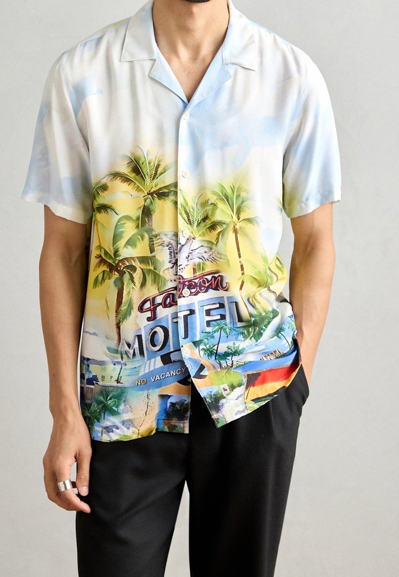 Camisa de manga corta con cuello que presenta un estampado tropical con palmeras, un letrero de motel y un fondo colorido. Hecha de tela ligera.