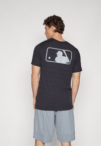 T-shirt de algodão azul-marinho com um gráfico do logo da MLB em branco nas costas, gola redonda e mangas curtas. Combinado com calções cinza-claro.