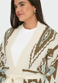 Ženska, oblečena v belo srajco z bež, modro in rjavo vzorčasto jopico, pripeto okoli pasu, prav tako nosi teksturirane zlate uhane v obliki obroča.