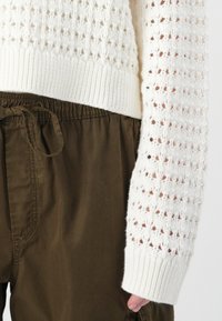 White knitted sweater with a textured pattern, featuring a ribbed hem and sleeves. Paired with dark olive brown elastic waist trousers.