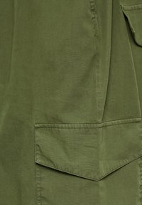 Olivenfarbener Stoff mit glatter Textur, ausgestattet mit einer großen aufgesetzten Tasche mit schrägem Klappverschluss und sauberem Stich entlang der Kanten.