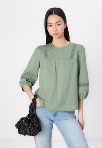 Blusa verde, con texture, a collo rotondo, maniche tre quarti e cuciture decorative; abbinata a jeans blu e una borsa nera con motivo.