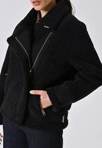 Giacca in pile nera con ampio colletto, chiusura con zip e tasche laterali. Presenta una finitura testurizzata, accessori in oro e un cinturino nero decorativo.