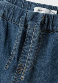 Pantaloni in denim con tonalità blu, vita elastica con cordini regolabili e dettagli di cucitura a contrasto. L'etichetta è visibile all'interno.
