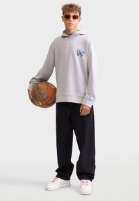 Felpa grigia con grafica sul petto, abbinata a pantaloni larghi neri. Tiene un pallone da basket consumato, indossando occhiali da sole colorati e scarpe bianche.