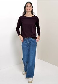 Top de manga larga color burdeos combinado con jeans de pierna ancha azul y zapatillas blancas, de pie frente a un fondo neutro.