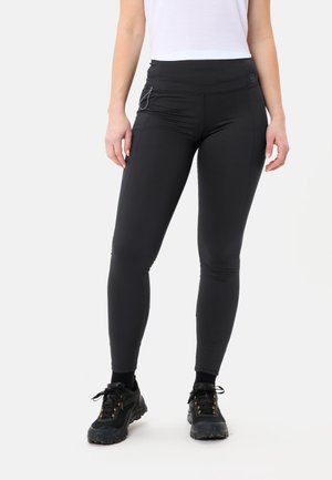 Schwarze Leggings mit hohem Bund, enger Passform und seitlichen Taschen. Getragen mit schwarzen Sportschuhen und einem weißen Oberteil. Dehnbares Material.