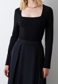 Schwarzes Langarmoberteil mit quadratischem Ausschnitt, kombiniert mit einem fließenden, plissierten schwarzen Rock. Sanfte Textur, minimalistisches Design.