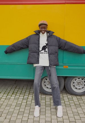 Veste noire gonflante avec des manches larges sur un pull blanc mettant en avant un graphique de kangourou, associé à un jean gris et des baskets roses.