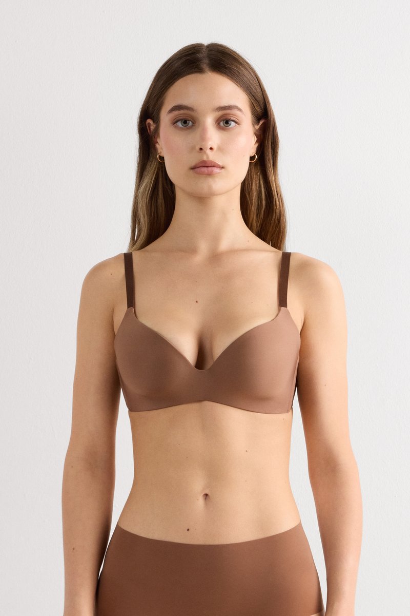 Femme aux longs cheveux bruns portant un soutien-gorge marron sans couture et une culotte taille haute assortie, debout devant un fond clair uni.