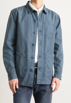 Mężczyzna w niebieskiej kurtce zapinanej na guziki z trzema przednimi kieszeniami, na białej koszuli i ciemnych dżinsach, stojący na jednolitym tle.