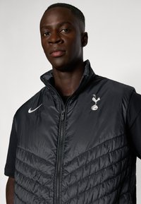 Schwarzte Pufferweste mit gestepptem Design und hohem Kragen. Verfügt über einen Reißverschluss, das Nike-Logo und das Emblem von Tottenham Hotspur auf der Brust.