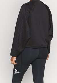 Sweat-shirt sportif noir avec manches et dos côtelés, arborant un détail de marque. Associé à un legging noir portant un logo blanc.