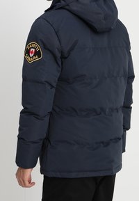 Mand iført en marineblå pufferjakke med hætte, der viser Zavetti Canada-patch på venstre overarm, står foran en ensfarvet baggrund.