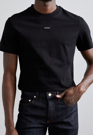 Man wearing a black short-sleeve t-shirt with small white "SANDRO" text and dark denim jeans with one hand in pocket.