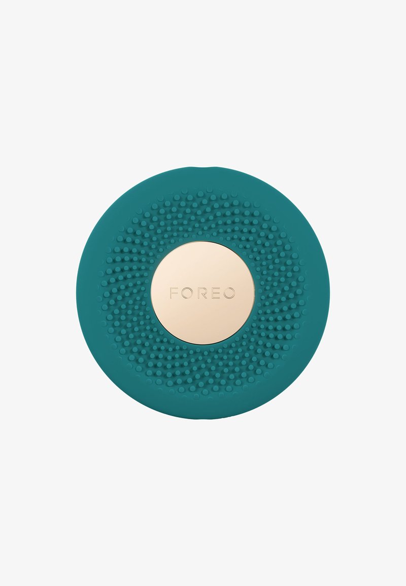 Teal silicone facial cleansing device with raised nodules and a gold circular center featuring the brand name "FOREO." Round, textured design.