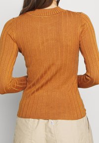 Rippstrick-Pullover in warmem Orangeton mit rundem Ausschnitt, figurbetonter Form und langen Ärmeln, der über ein strukturiertes Gewebedetail verfügt.