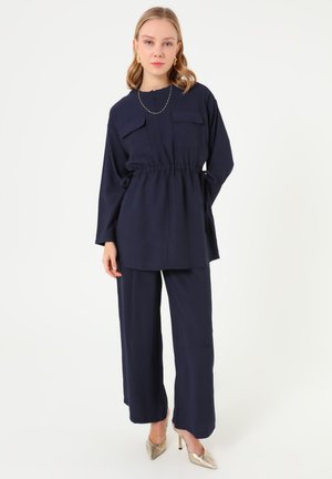 Woman wearing a navy blue long-sleeve tunic with drawstring waist and matching wide-leg pants, gold earrings, necklace, and metallic heels.