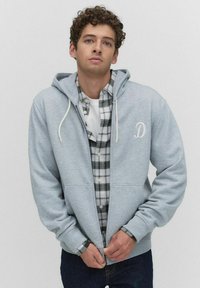 Jeune homme aux cheveux bouclés portant un sweat à capuche zippé gris clair sur une chemise à carreaux noir et blanc, en train de fermer la fermeture éclair du sweat.