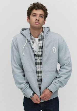 Jeune homme aux cheveux bouclés portant un sweat à capuche zippé gris clair sur une chemise à carreaux noir et blanc, en train de fermer la fermeture éclair du sweat.