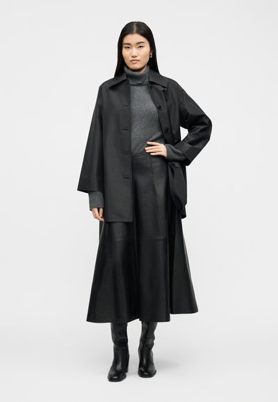 Γυναίκα στέκεται φορώντας μαύρο παλτό με κουμπιά, γκρι πουλόβερ ζιβάγκο, μαύρη δερμάτινη midi φούστα και μαύρες μπότες με τακούνι σε απλό φόντο.