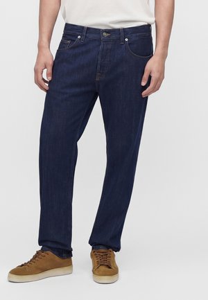 Mand iført mørkeblå jeans med løs pasform og brune ruskindssneakers med hvide såler, stående mod en ensfarvet lys baggrund.