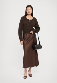 Brown knitted cardigan with rounded neckline, paired with a brown satin maxi dress. Black cylindrical handbag with shoulder strap and beige shoes.