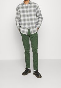 Mężczyzna w szarej i białej kratowanej koszuli noszonej na białym t-shircie, zielonych spodniach i czarnych butach, stojący z rękami w kieszeniach na jednolitym tle.