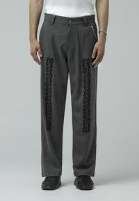 Gray plaid trousers with black decorative patterns along the sides, featuring a button closure and chain accent. Material is textured.