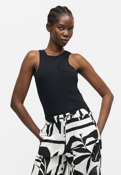 Γυναίκα με πλεξούδες στα μαλλιά, που φοράει μαύρο αμάνικο τοπ και ψηλόμεσο λευκό παντελόνι με έντονα μαύρα μοτίβα φύλλων.