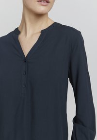 Blusa azul marino con escote en V, tres botones y mangas largas de textura suave y lisa. Diseño simple y clásico.