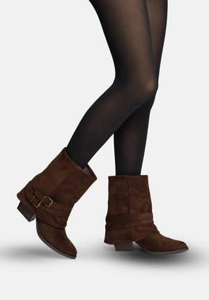 Gambe in collant neri che indossano stivali marroni in camoscio fino a metà polpaccio con fibbie doppie e tacchi bassi a blocchetto su uno sfondo semplice.