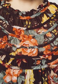 Blouse fleurie en tissu foncé avec des volants superposés, ornée de motifs floraux orange, jaune et vert, avec des accents bleu clair le long des bords.