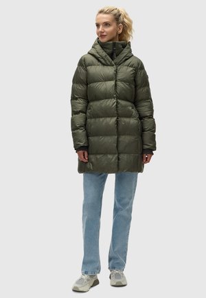 Vadderad, olivgrön pufferjacka med hög krage och huva, med horisontell sömnad och dragkedja fram. Bärs över ljusblå jeans.