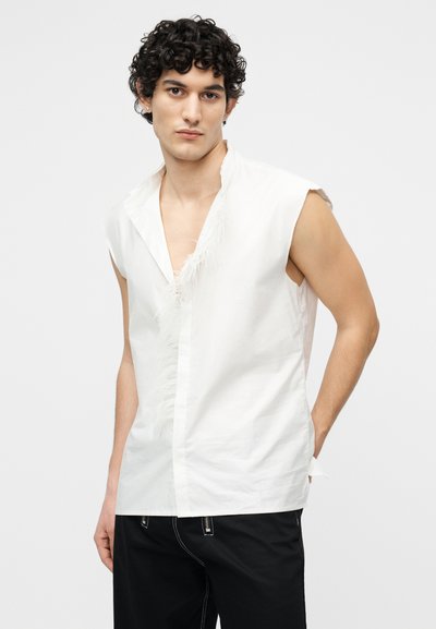 Νέος άνδρας με σγουρά μαλλιά που φοράει άσπρο αμάνικο πουκάμισο με σκισμένες άκρες και μαύρο παντελόνι, στέκεται μπροστά σε απλό φόντο.