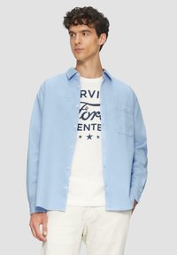 Helle blaue Hemdbluse aus einem texturierten Stoff, mit einer Brusttasche, über einem weißen T-Shirt mit schwarzen Grafiken getragen.