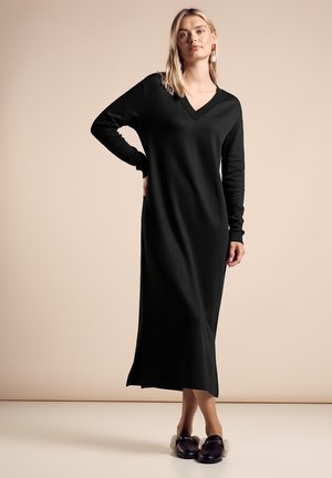 Svart långärmad klänning med V-ringning i en åtsittande design, tillverkad av ett slätt tyg med en knä-hög slits, kombinerad med mörka loafers.