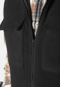 Schwarze Fleece-Weste mit einem Frontreißverschluss, zwei Brusttaschen und einem gemusterten Hemd darunter in Orange- und Grautönen.