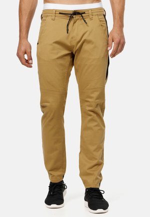Herrenbeige Freizeithose mit Kordelzug in der Taille, seitlichen Taschen und schwarzen Sneakers vor weißem Hintergrund.