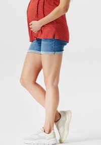 Červený top s květinovým vzorem, modré denimové šortky s ohrnutými nohavicemi, bílé tenisky s texturovanými detaily. Důraz na nohy a trup.