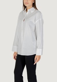 Camicia bianca con colletto button-up, caratterizzata da una sottile trama a disegno, maniche lunghe e polsini a contrasto. Abbinata a pantaloni neri aderenti.