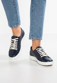 Marina läderskor med krämfärgade snören, med rund tå och vit sula. Bärs tillsammans med blå denimjeans, som är något avkortade vid ankeln.