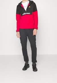 Sudadera con capucha roja y negra con cremallera frontal, logo "ellesse" en el pecho; combinada con pantalones cargo negros y zapatillas negras.