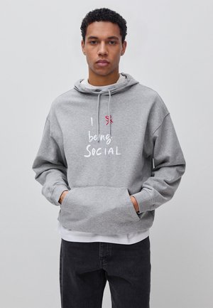 Joven con cabello rizado que lleva una sudadera gris con el texto "I love being social", de pie frente a un fondo claro y liso.