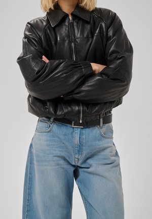 Personne portant une veste en cuir noire zippée avec des poignets élastiques et un jean taille haute bleu clair, les bras croisés sur la poitrine.