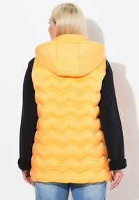 Gelbe gepolsterte Weste mit Kapuze, die ein Zickzack-Steppmuster auf dem Rücken aufweist, kombiniert mit einem schwarzen Langarmoberteil und blauen Jeans.