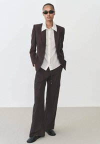 Donkerbruine blazer met scherpe revers over een wit gestreept shirt. Losvallende donkere broek met zakken en een koord, gecombineerd met zwarte schoenen.