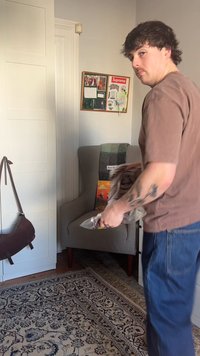 Mies, jolla on tatuointi oikeassa kyynärvarressa, pitää muovilla käärittyä esinettä olohuoneessa, nojatuolin vieressä, jonka päällä on viltti ja seinäkollaasi.