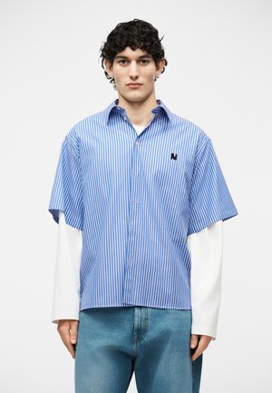 Jeune homme aux cheveux bouclés portant une chemise à manches courtes bleu et blanc à rayures verticales par-dessus une chemise blanche à manches longues et un jean bleu.